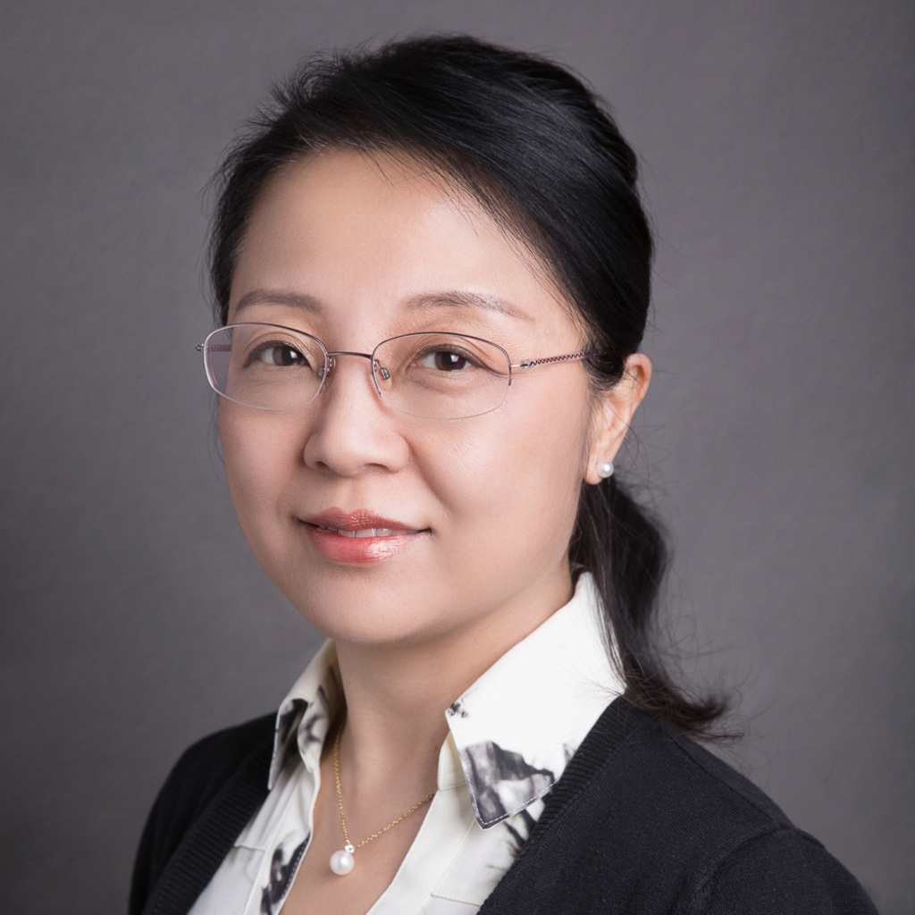 A person wearing a black cardigan, patterned white shirt, and pearl necklace, against a gray background.
