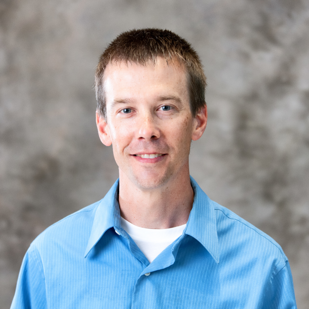 A person wearing a blue collared shirt and white undershirt, standing in front of a gray background.