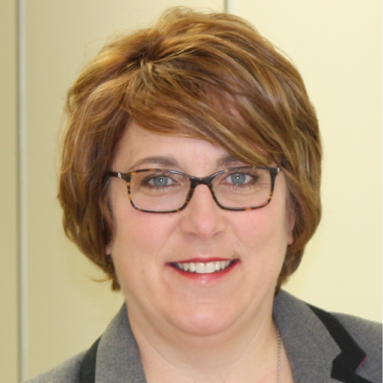 Person with short light brown hair and glasses, smiling, wearing a grey blazer and light top against a plain background.