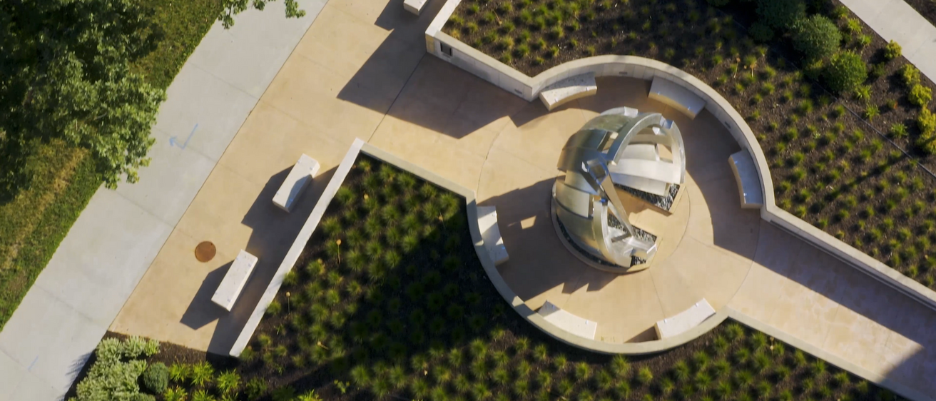 Aerial photograph of a sculpture composed of interconnected metal rings and panels, situated within a circular courtyard with concrete benches. The courtyard is bordered by paved paths and green landscaping with shrubs and grasses.