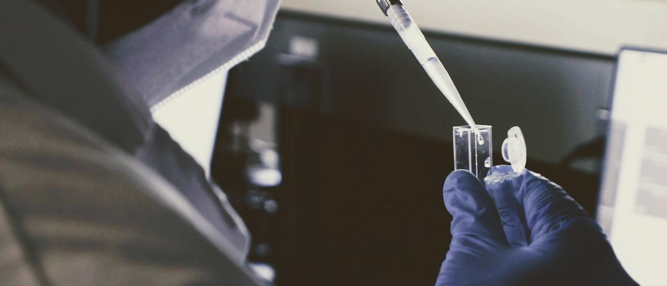 A person wearing protective clothing and gloves is using a pipette to transfer liquid into a small transparent container in a laboratory setting. The background includes laboratory equipment and a computer screen.