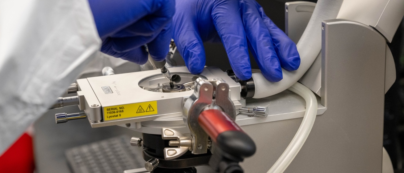 Close-up of gloved hands operating a freeze-drying microscope in a laboratory, adjusting the sample holder to analyze liquid crystallization properties.
