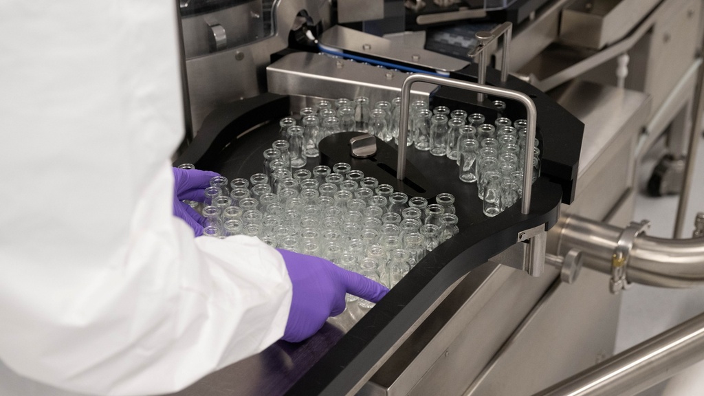 Person in lab coat and gloves handling glass vials on a conveyor in a lab setting.