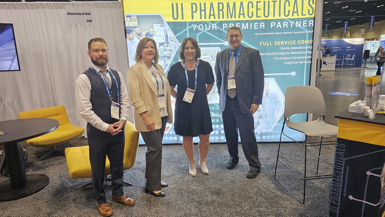 A trade show booth for UI Pharmaceuticals (University of Iowa) displaying banner text "YOUR PREMIER PARTNER" and services including sterile manufacturing and formulation development. Four professionally dressed staff members wearing conference badges stand in front of the booth, which features yellow seating and University of Iowa branding.
